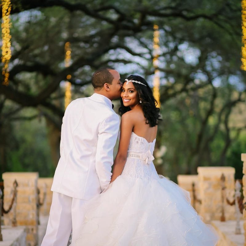 groom kisses bride on temple as she smiles at camera under canopy of trees